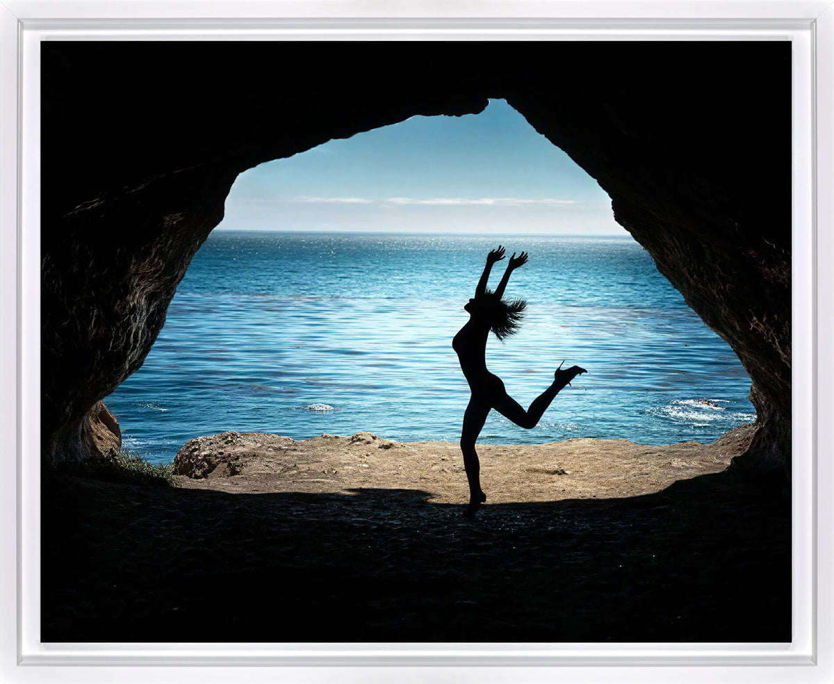 A framed photographic print showing a silhouetted dancer jumping in front of a cave opening that reveals a bright blue ocean and sky.