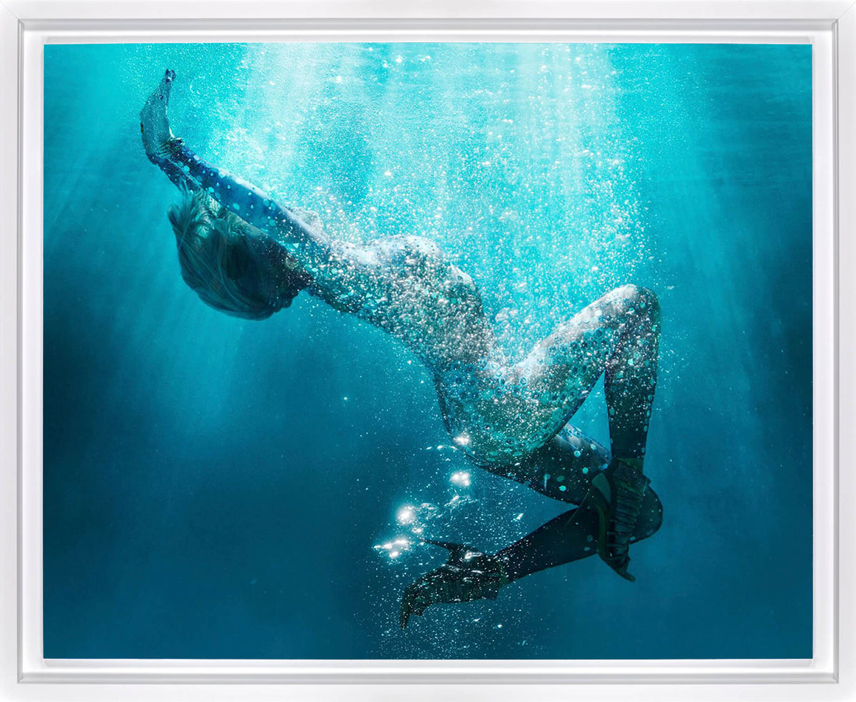 Underwater art print featuring a woman diving downward through bright turquoise blue water with rising bubbles and light rays filtering from above, presented with a white frame.